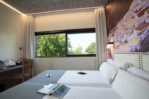 A hotel room with a bed, a desk with a laptop, a chair, and a window with a view of trees.