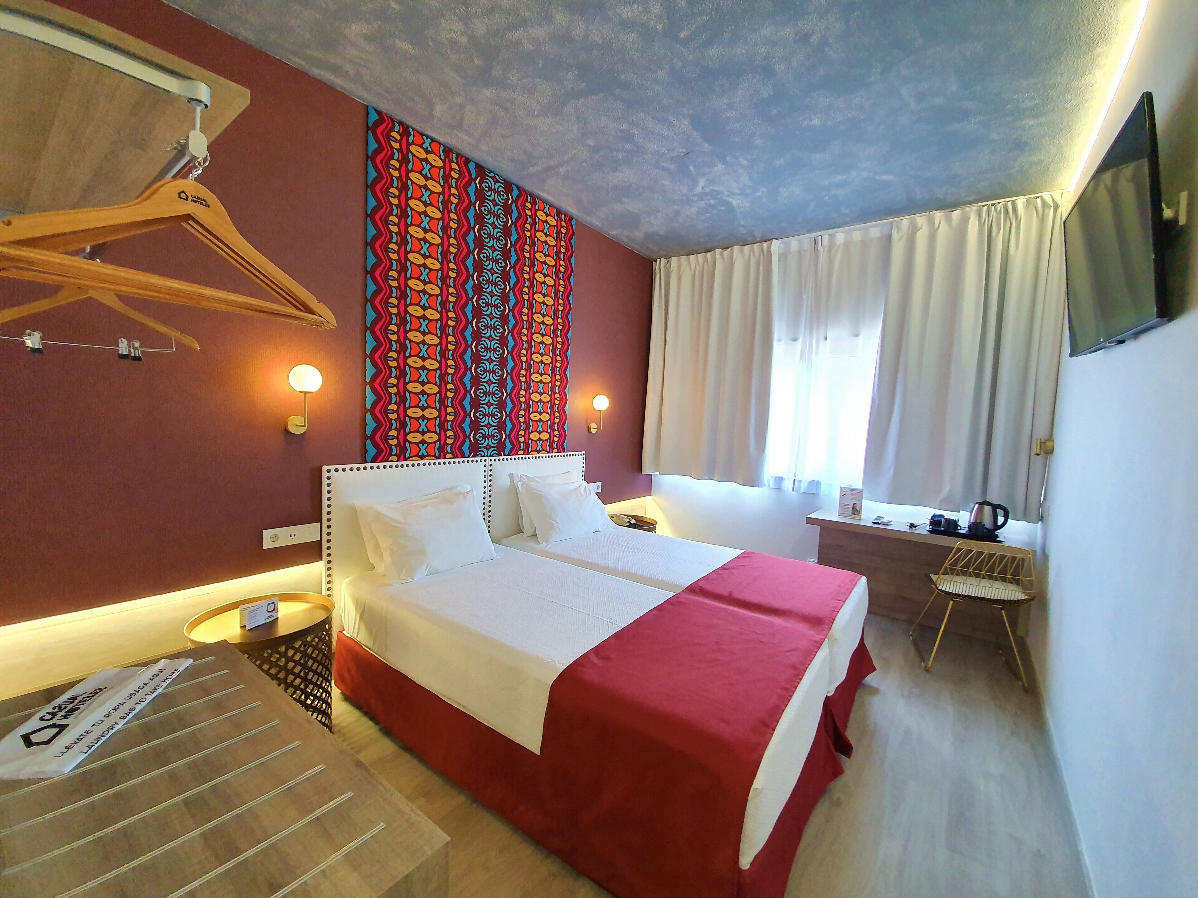 A hotel room with a bed, bedside tables, a desk, a TV, and a window with curtains.