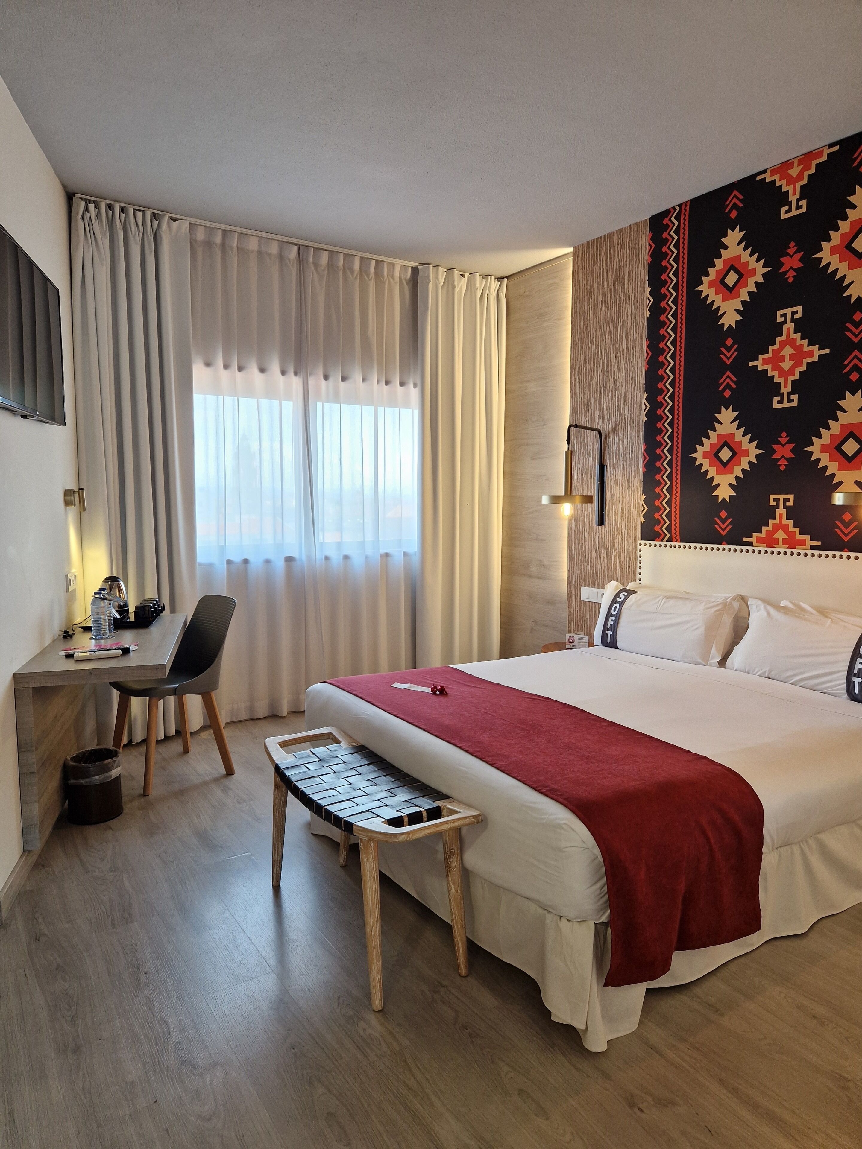 A hotel room with a bed, a desk with a chair, a small table, and a window with curtains.