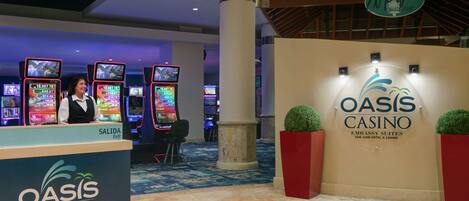Zona de recepción de un casino, con un cartel que indica Oasis Casino, una ventanilla de caja y varias máquinas tragamonedas.