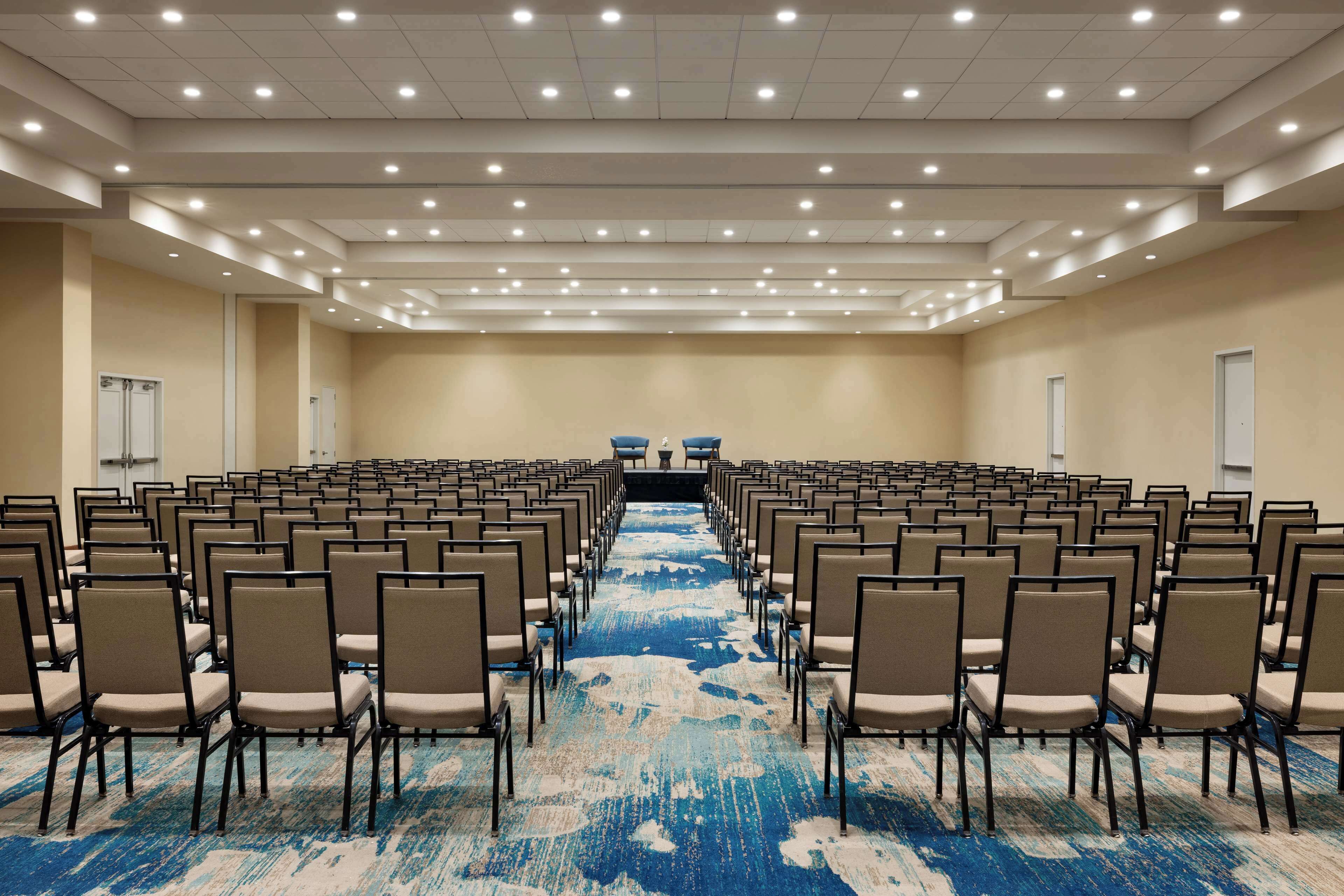A large conference room with rows of chairs facing a stage, beige walls, and a blue carpet.