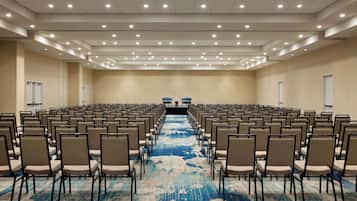 A large conference room with rows of chairs facing a stage, beige walls, and a blue carpet.