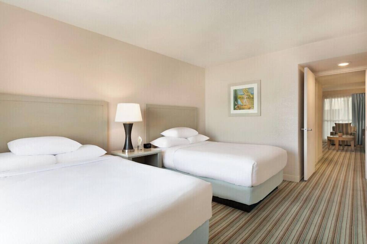 Quarto de hotel com duas camas, uma mesa de cabeceira com abajur, um quadro na parede e vista para uma sala de jantar através de uma porta aberta.