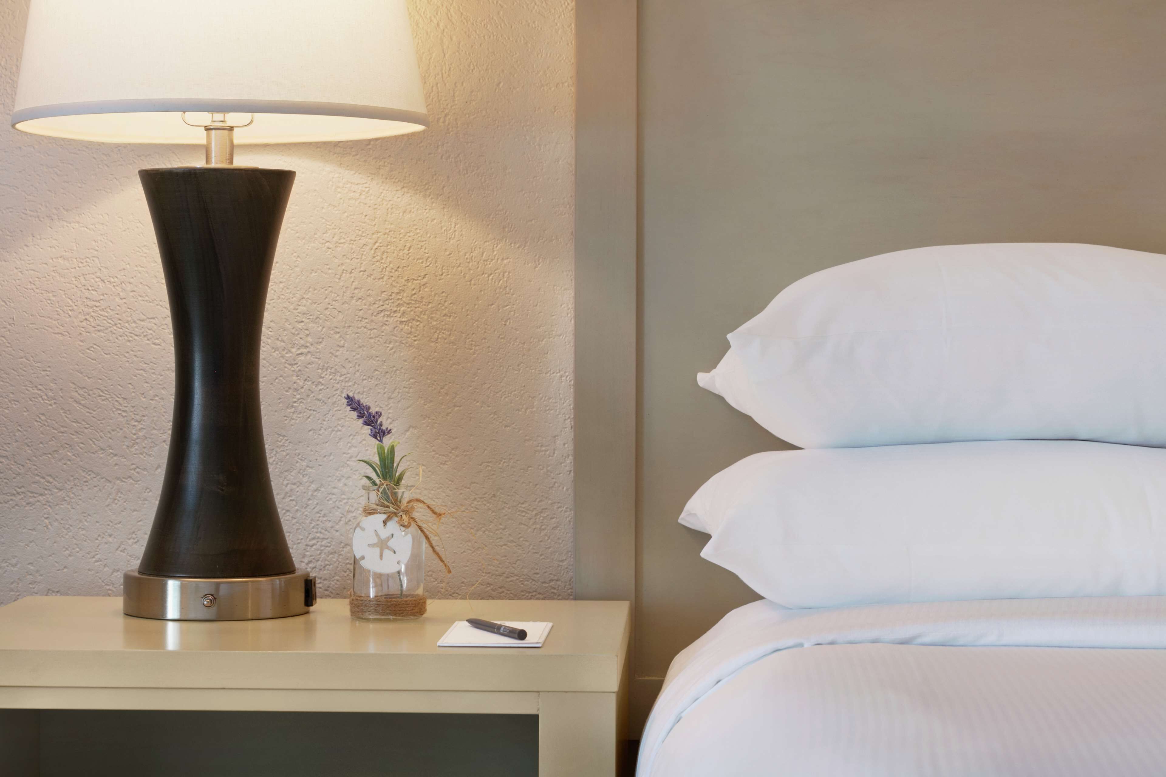 A neatly made bed with white pillows and a bedside table with a lamp, a remote control, and a small vase with flowers.
