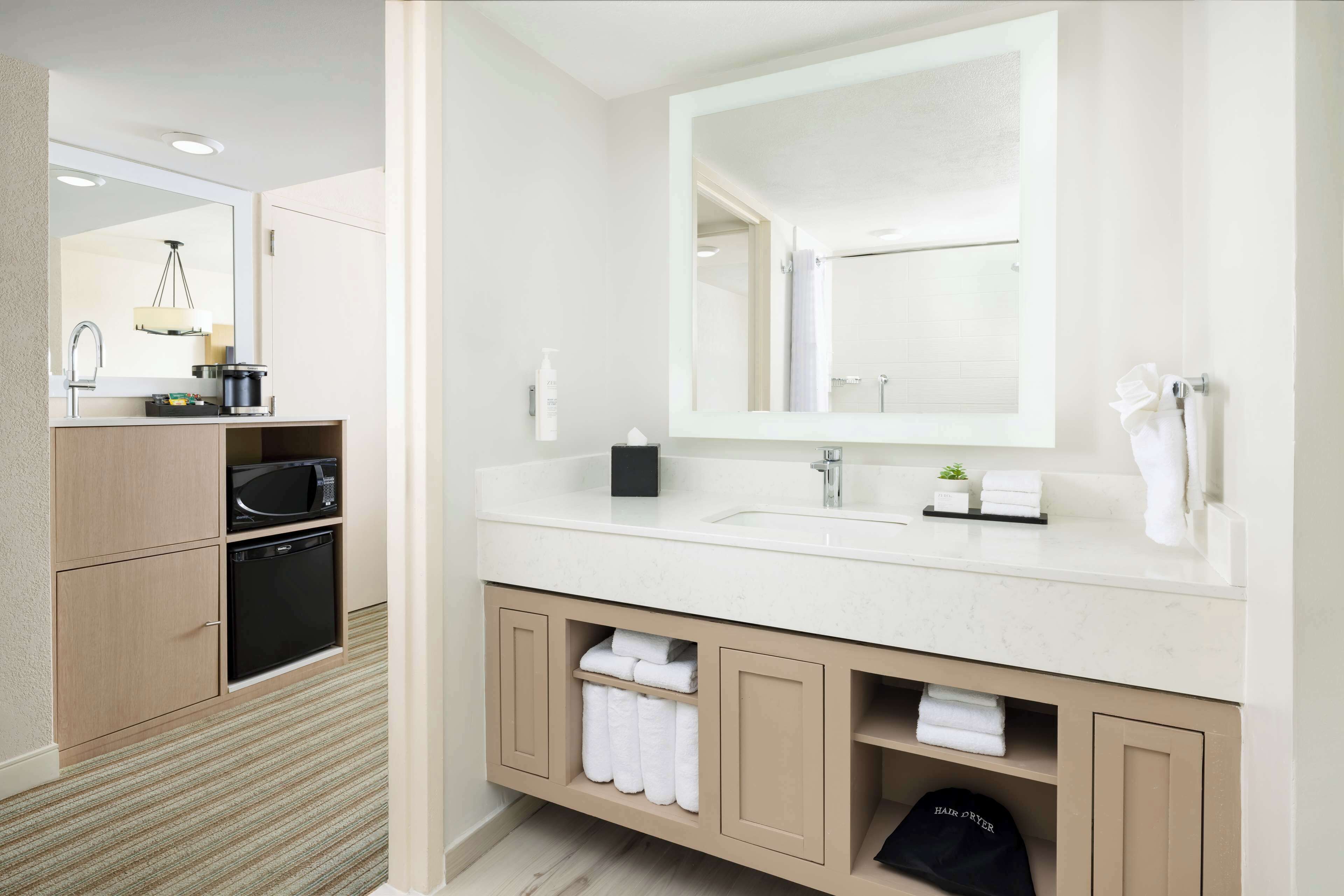 Un baño moderno con un espejo grande, un lavamanos y un microondas. Se ven toallas y artículos de aseo.