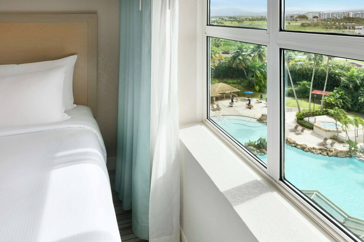 Quarto de hotel com cama, janela e vista para piscina e área externa.