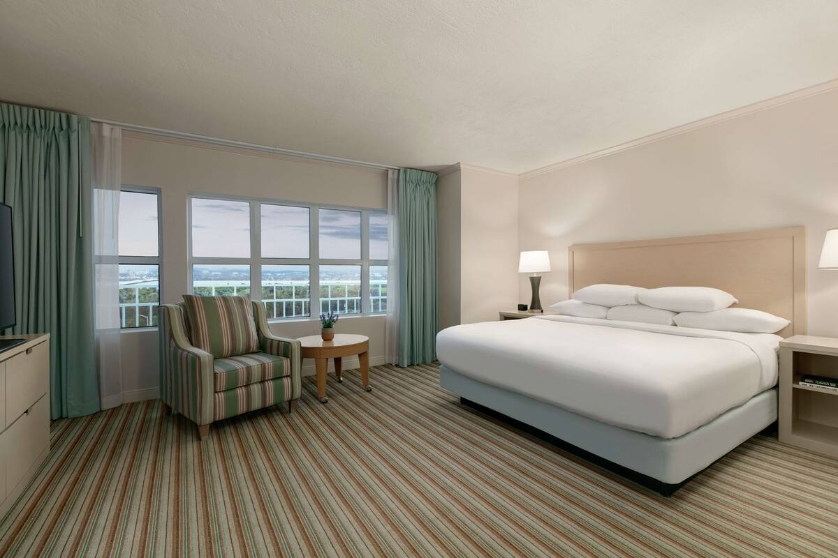 Quarto de hotel com uma cama grande, uma cadeira, uma mesinha e uma televisão.