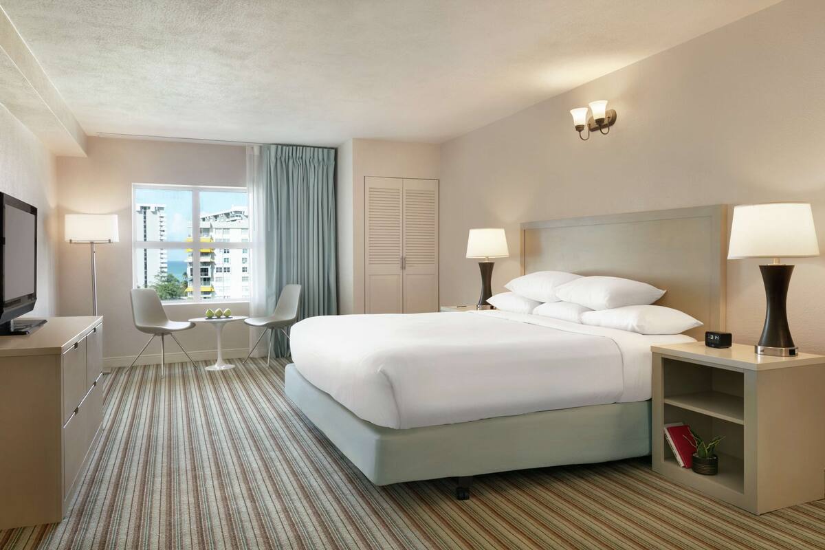 Quarto de hotel com uma cama grande, duas mesas de cabeceira com luminárias, uma TV, uma pequena mesa com uma cadeira e uma janela com cortinas.