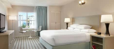 Habitación de hotel con una cama grande, dos mesitas de noche con lámparas, un televisor, una pequeña mesa con una silla y un ventanal con cortinas.