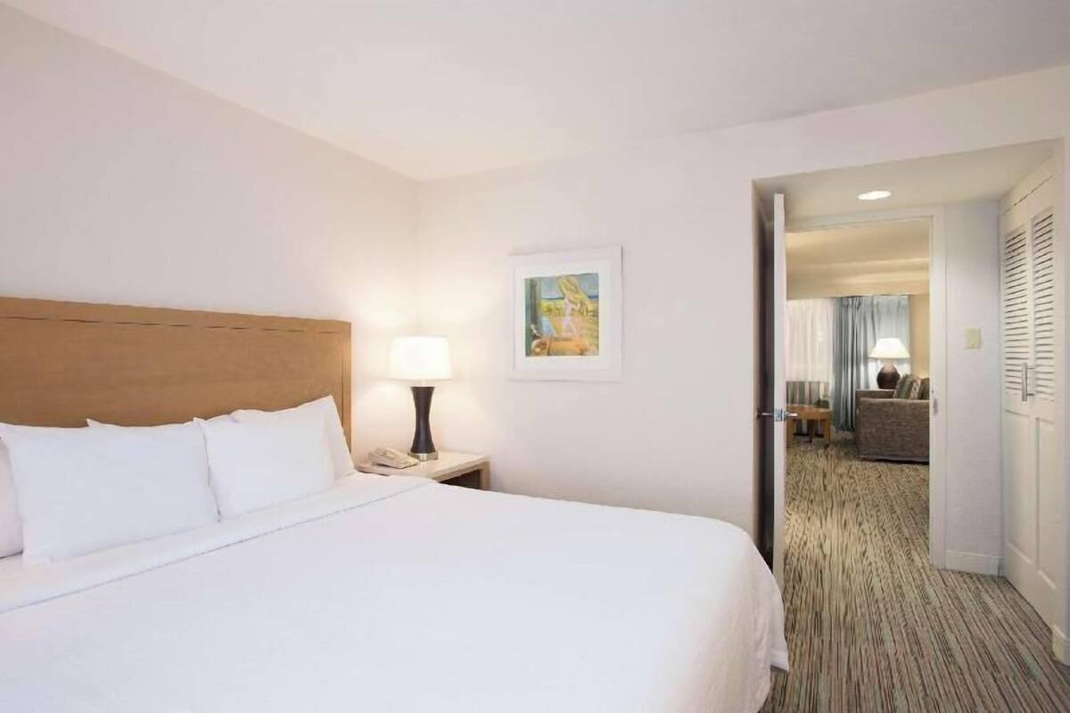 Quarto de hotel com cama, mesa de cabeceira com abajur, quadro na parede e vista para a sala de estar adjacente.