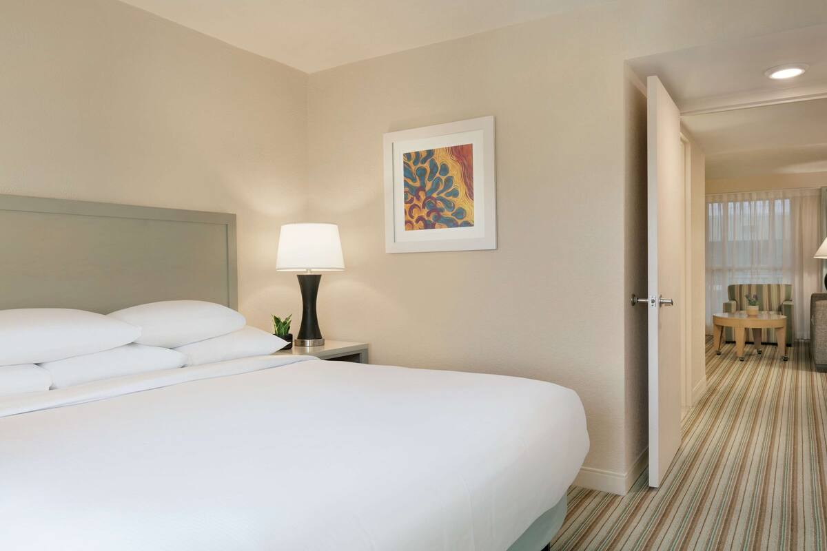 Quarto de hotel com uma cama grande, mesa de cabeceira com luminária, obra de arte emoldurada na parede e porta que dá para uma área de estar.
