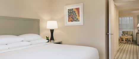 Un cuarto de hotel con una cama grande, una mesita de noche con lámpara, una obra de arte enmarcada en la pared y una puerta que da a un área de descanso.