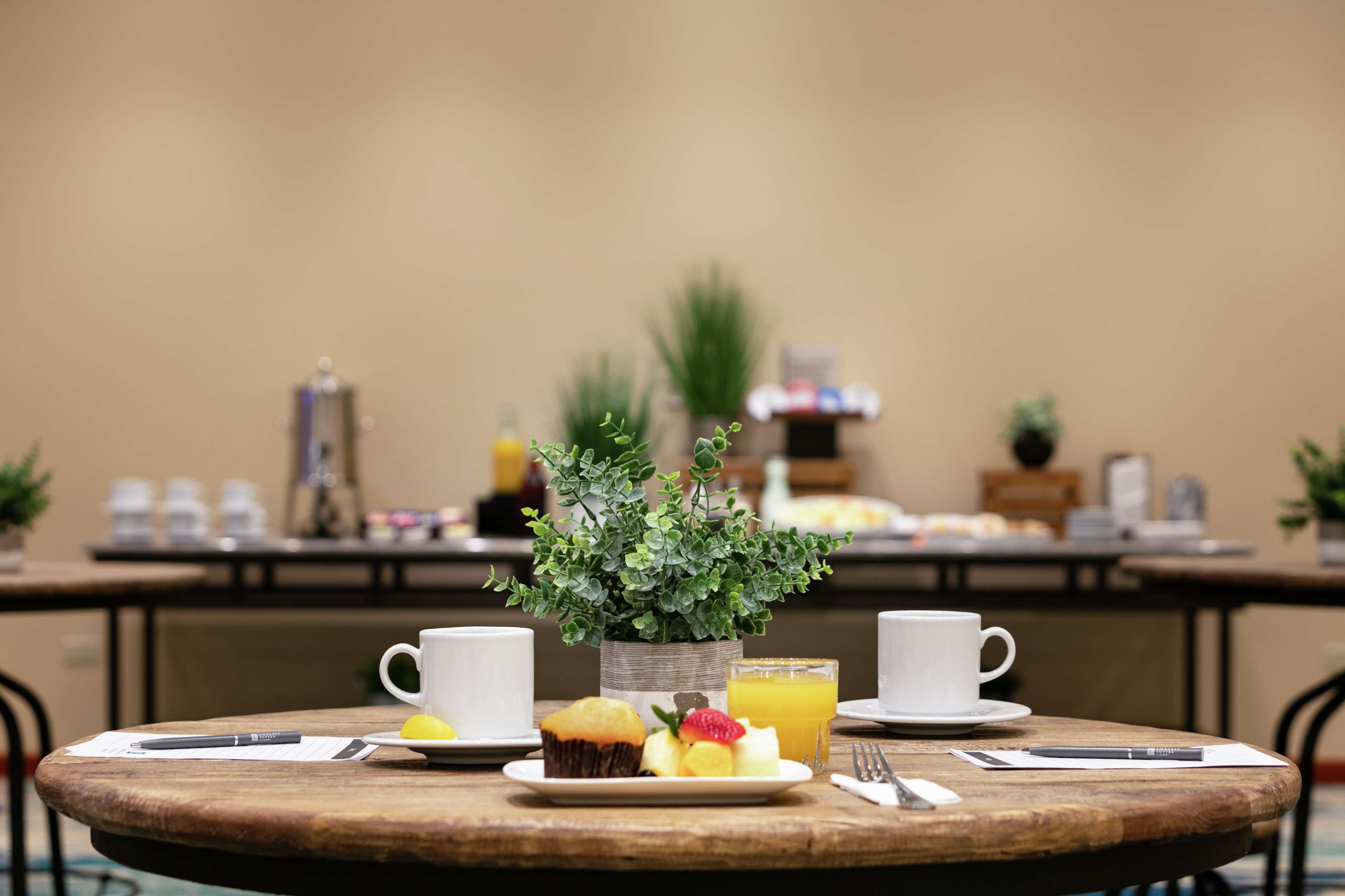 Una mesa redonda de madera con una taza blanca, un plato con un muffin y fruta, un vaso de zumo de naranja y una pequeña planta en maceta.