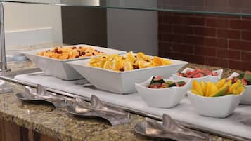 A buffet with various fruit options in white bowls, arranged on a granite countertop with metal serving utensils.