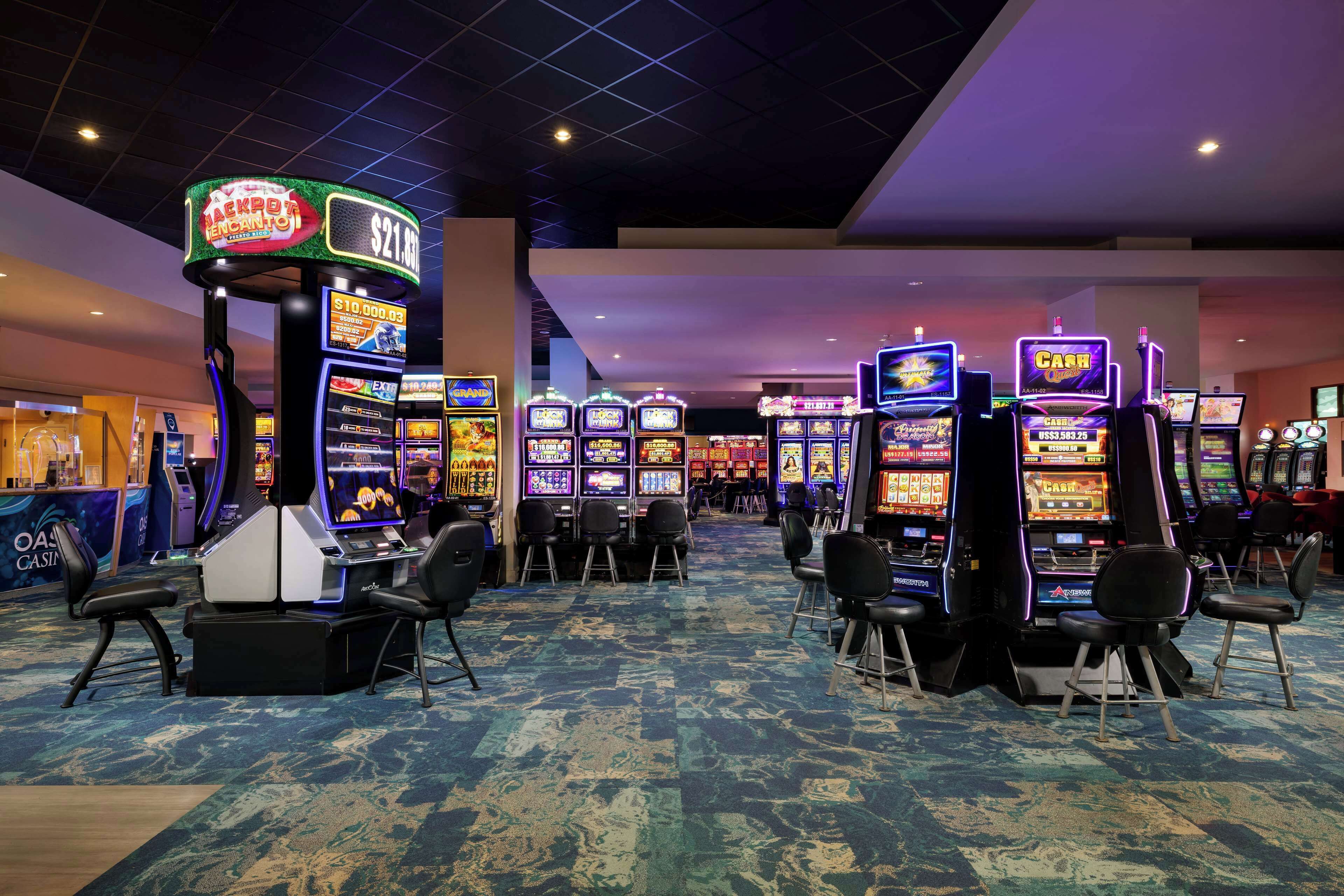 A casino floor with multiple slot machines, chairs, and a carpeted floor.