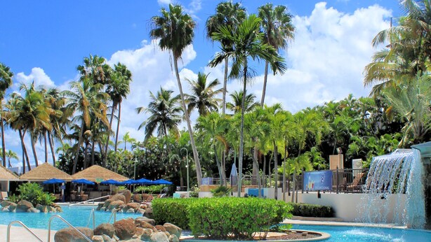 A resort pool with palm trees, a waterfall, and lounge areas. - Embassy Suites by Hilton San Juan Hotel & Casino (Carolina)