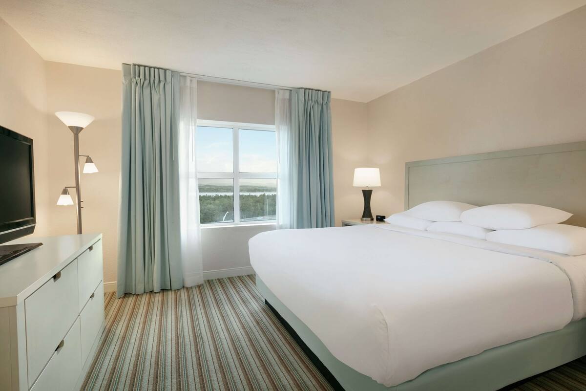 Quarto de hotel com uma cama grande, uma televisão em uma cômoda, um abajur e uma janela com cortinas.
