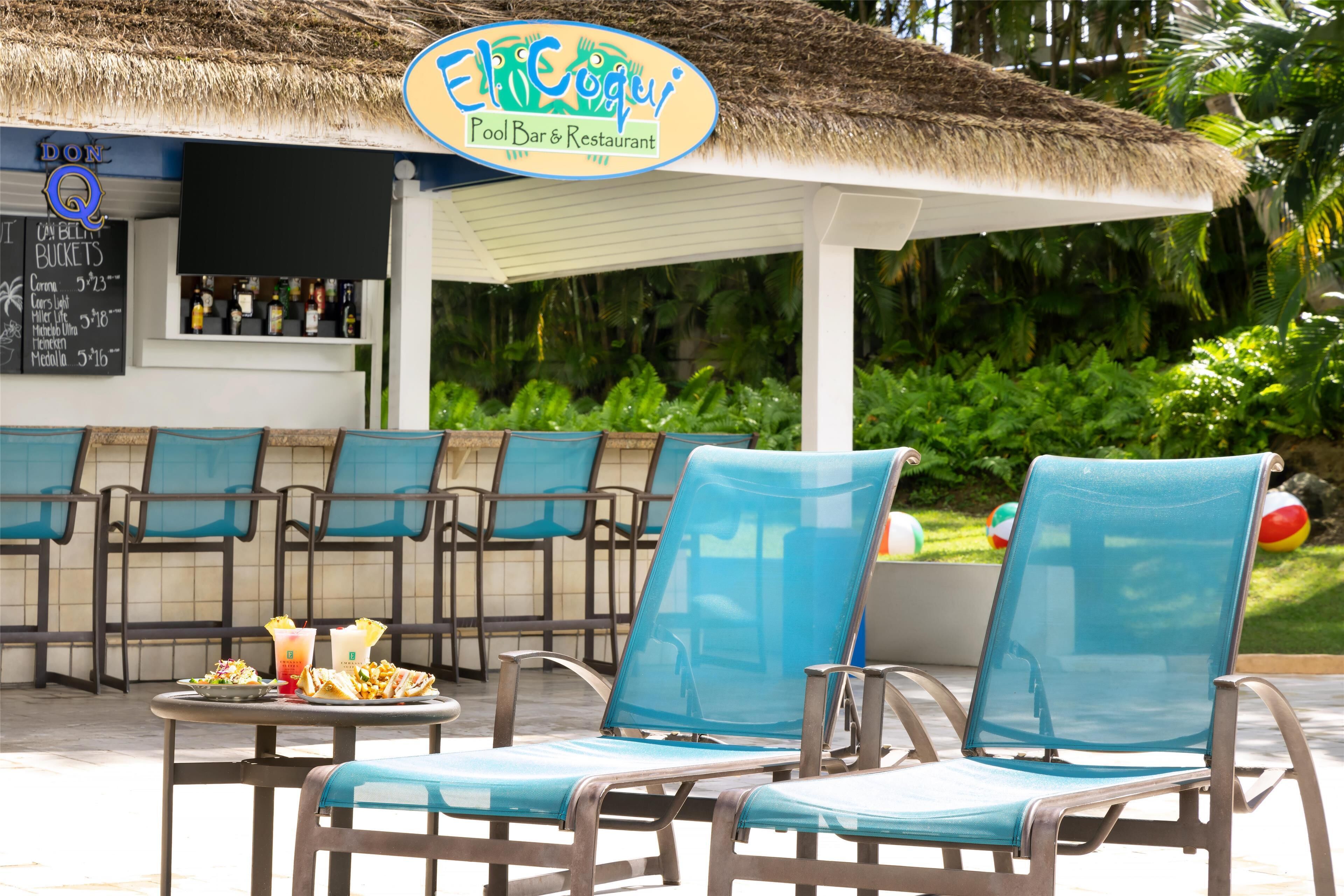 A poolside area with blue cushioned chairs, a table with food and drinks, and a bar with a sign reading "El Coguai Pool Bar & Restaurant".