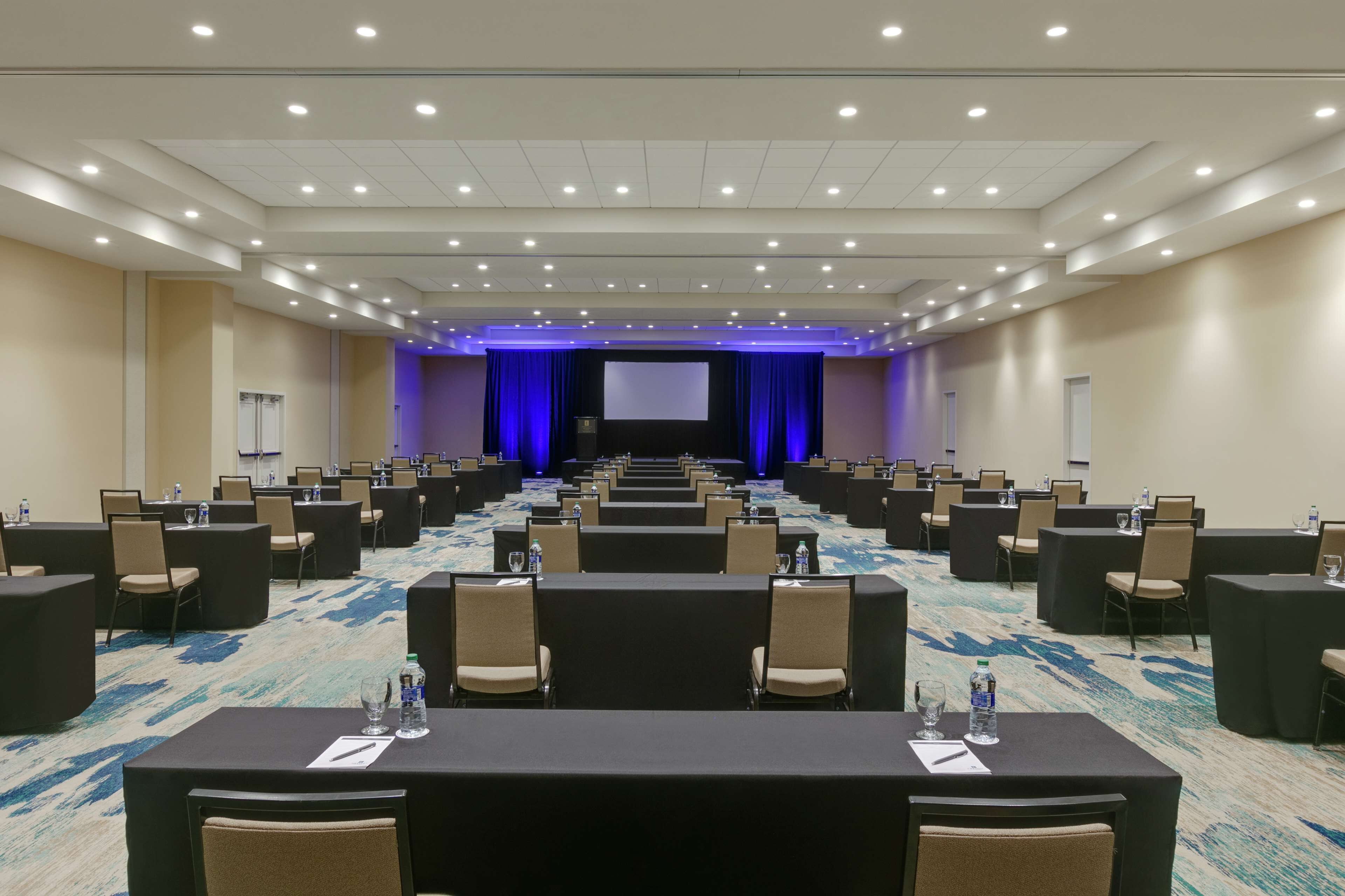 A conference room with rows of tables and chairs, a large screen at the front, and a carpeted floor.