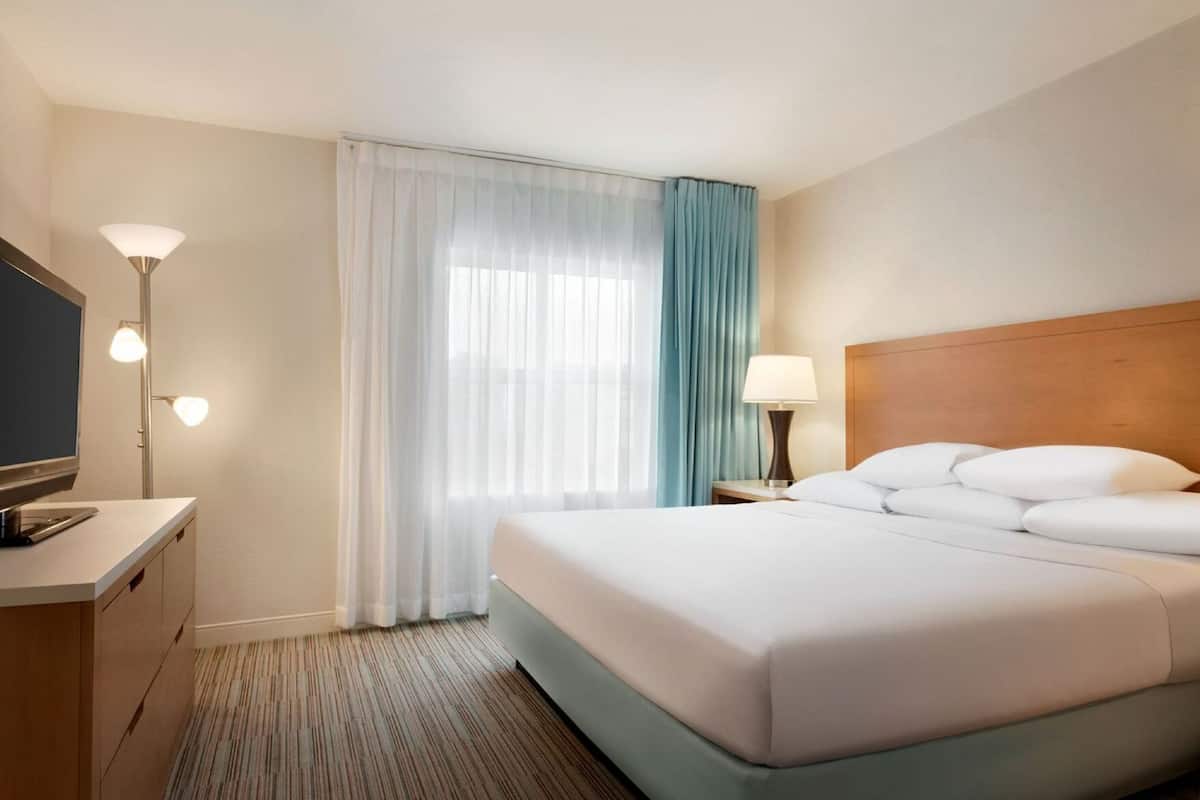 Quarto de hotel com cama, televisão, luminária, mesa de cabeceira e janela com cortinas.