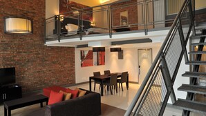 Ein geräumiger Wohnbereich mit hohen Decken, einer Backsteinwand, einem Esstisch mit Stühlen und einer Treppe mit Metallgeländer.