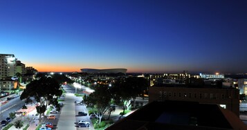 Une vue d’une ville au crépuscule, avec un stade imposant et une rue animée, bordée d’arbres et traversée par des voitures.