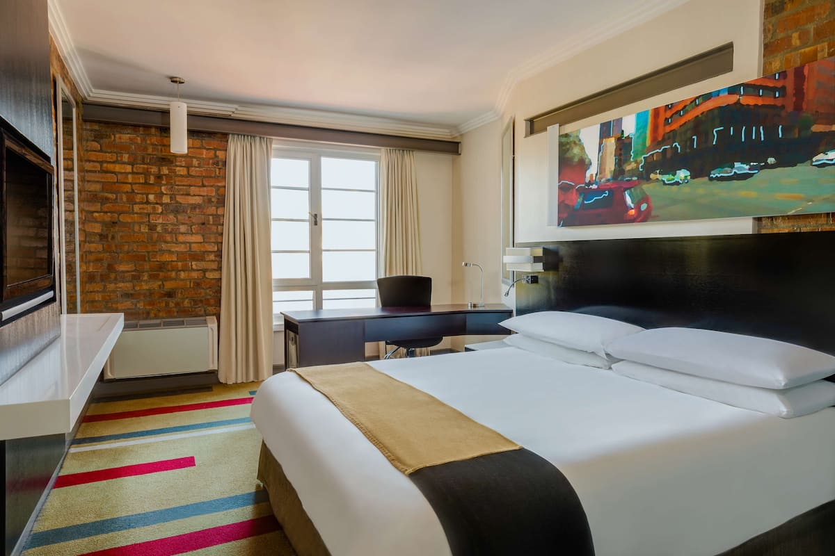A hotel room with a large bed, a desk, a chair, a TV, and a painting on the wall.