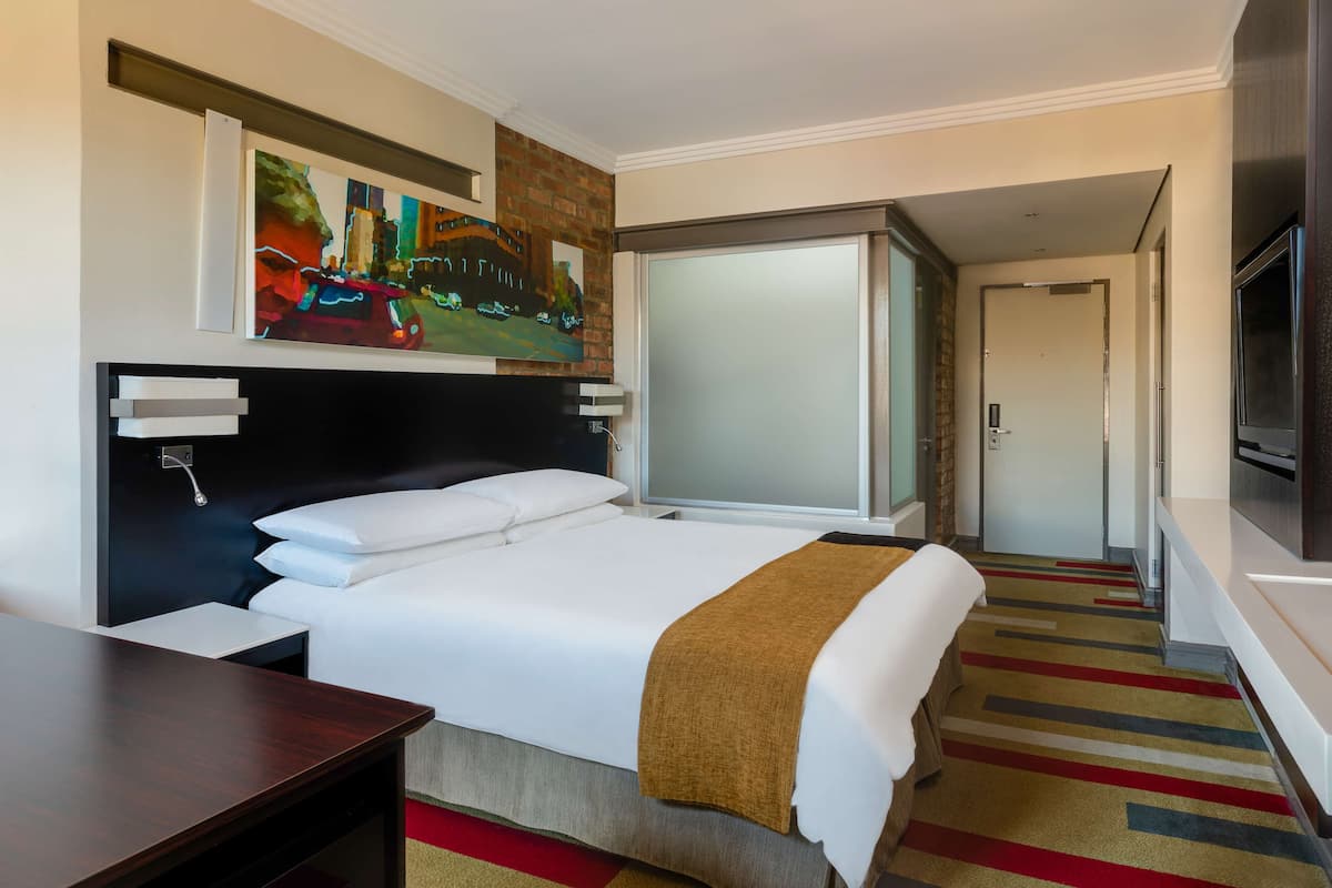 A hotel room with a bed, a desk, a TV, a painting, and a door leading to another room.