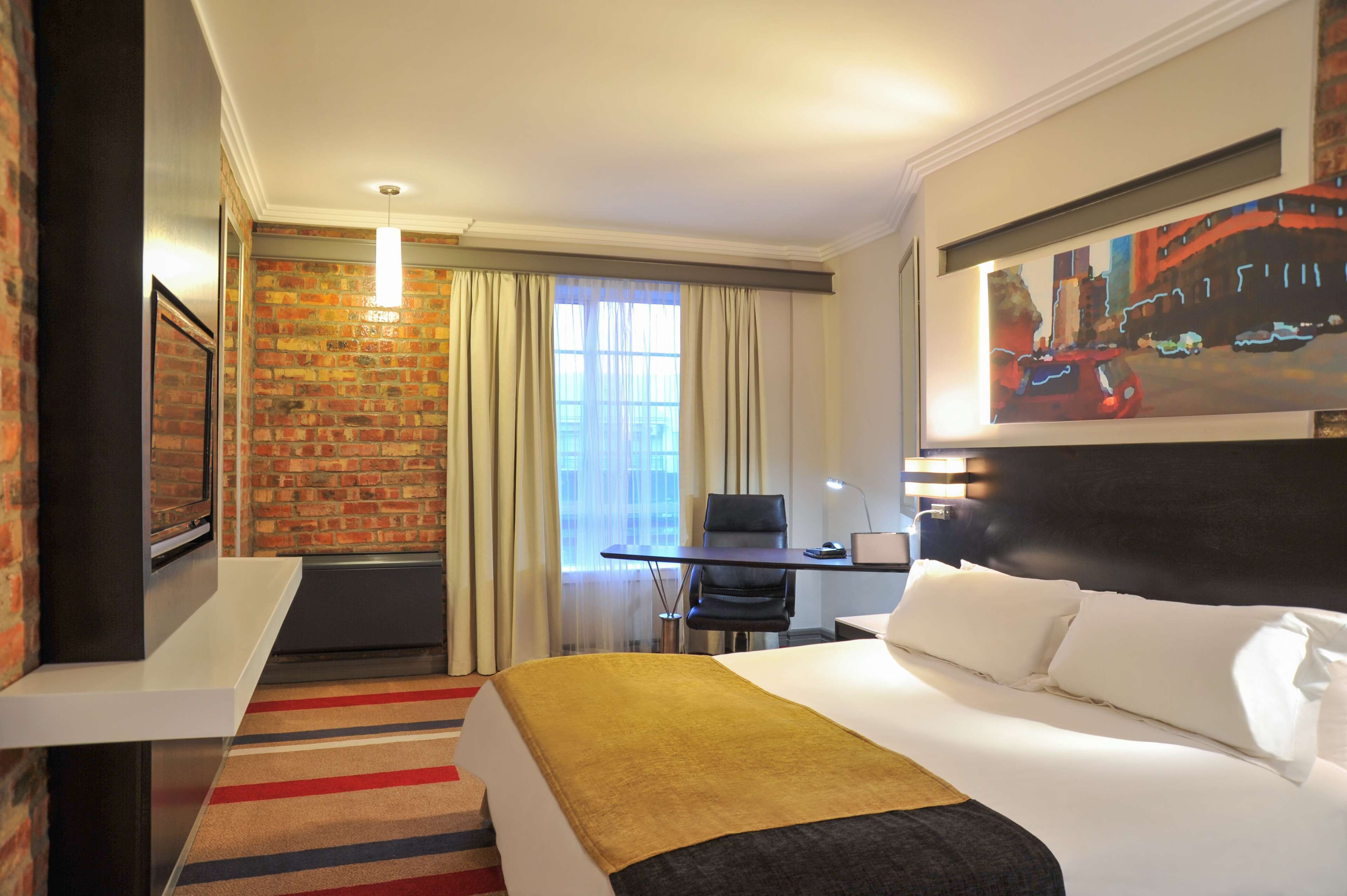 Habitación de hotel con cama, escritorio, silla, ventana con cortinas y un cuadro en la pared.