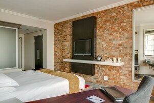 A hotel room with a brick wall, a flat-screen TV mounted above a fireplace, a bed with white bedding, and a desk with a chair.