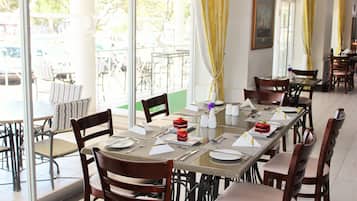 A restaurant with a long table set for a meal, chairs around it, and a view of the outside through large windows.