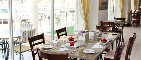 A restaurant with a long table set for a meal, chairs around it, and a view of the outside through large windows.