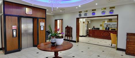 Un vestíbulo de hotel con una recepción, una araña y un mostrador de madera, además de una mesa con un florero.