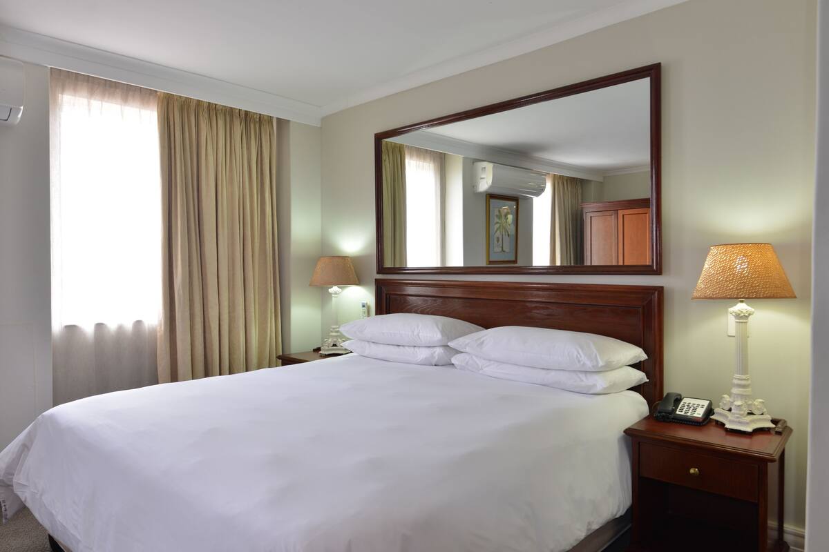 A hotel room with a large bed, a nightstand with a lamp, a mirror, and a window with curtains.