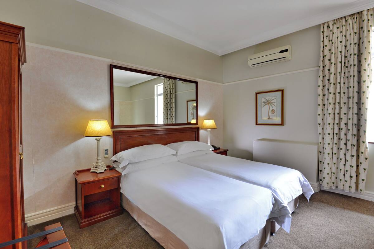 A hotel room with two beds, a large mirror, a nightstand with lamps, a framed picture, and a window with curtains.