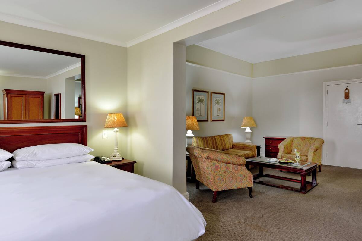 A hotel room with a large bed, two chairs, a small table, and a desk. There are lamps on both sides of the bed and a mirror above it.