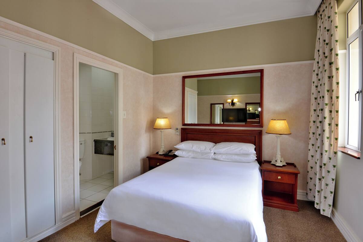 A hotel room with a bed, two bedside tables with lamps, a mirror above the bed, a wardrobe, and a bathroom visible through an open door.