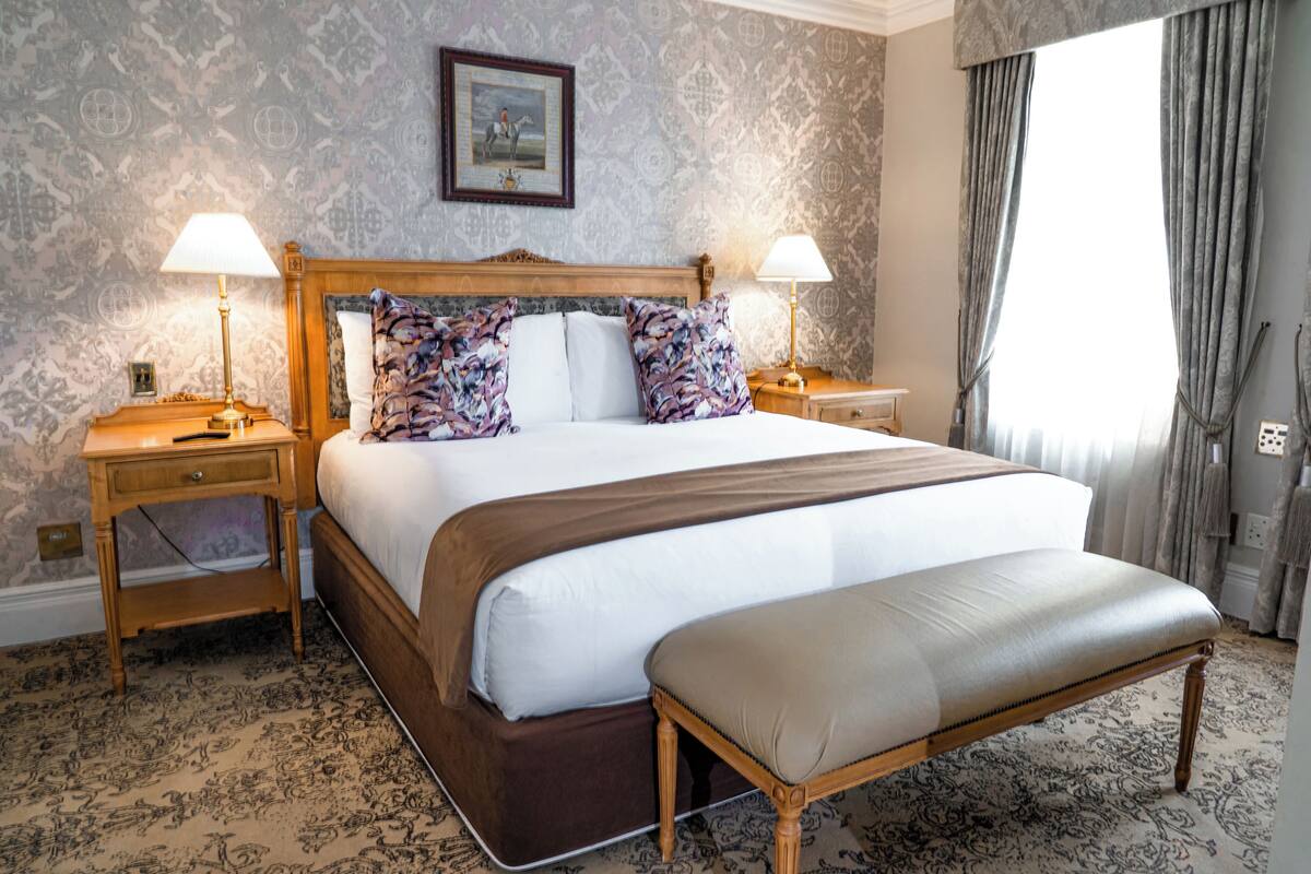 A large bed with a wooden headboard, two bedside tables with lamps, a bench, patterned wallpaper, and a framed picture on the wall.