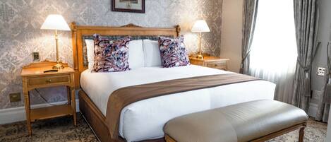 A large bed with a wooden headboard, two bedside tables with lamps, a bench, patterned wallpaper, and a framed picture on the wall.
