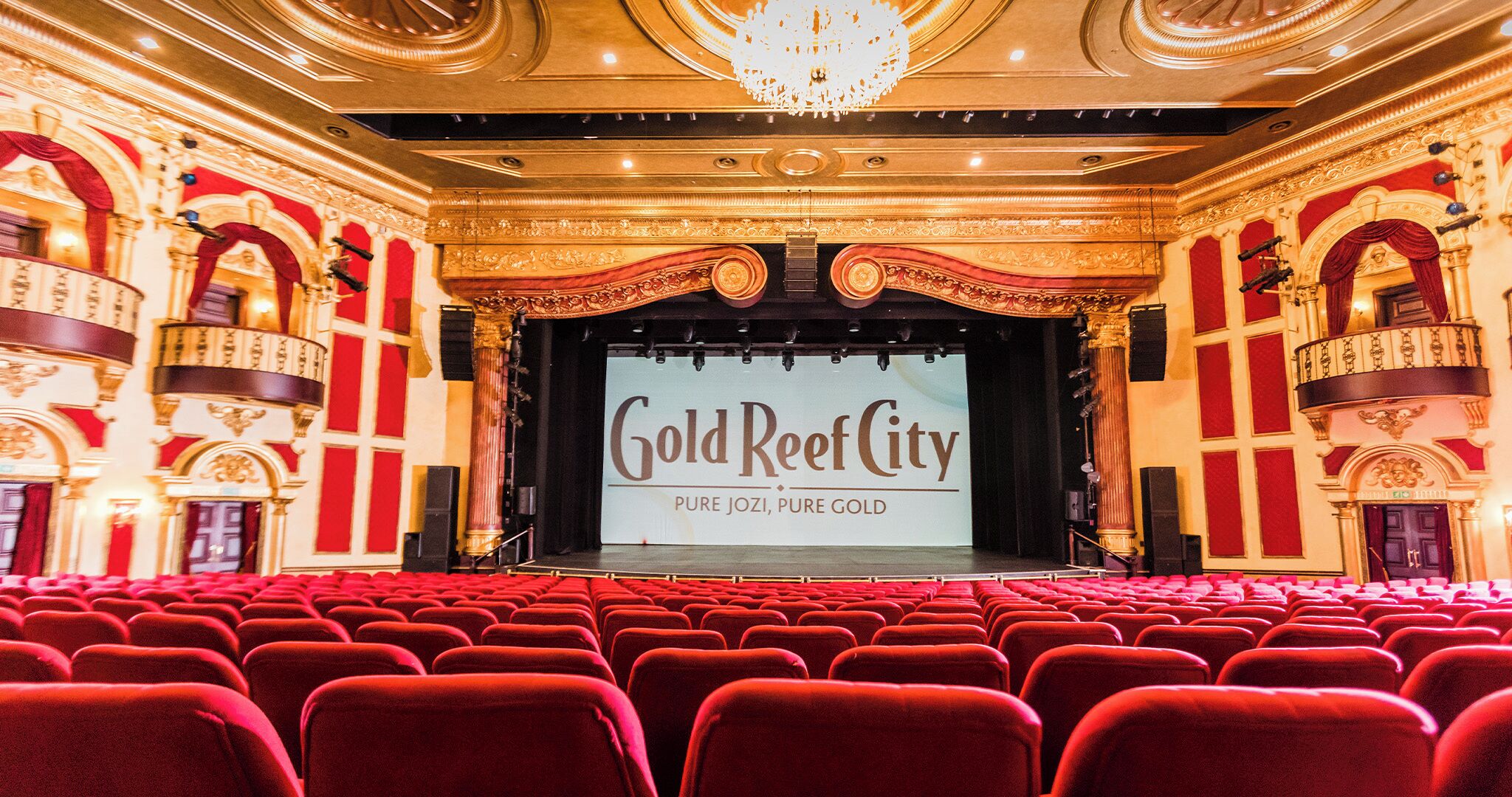 Un teatro con asientos rojos y un escenario que muestra el letrero de 'Gold Reef City'.