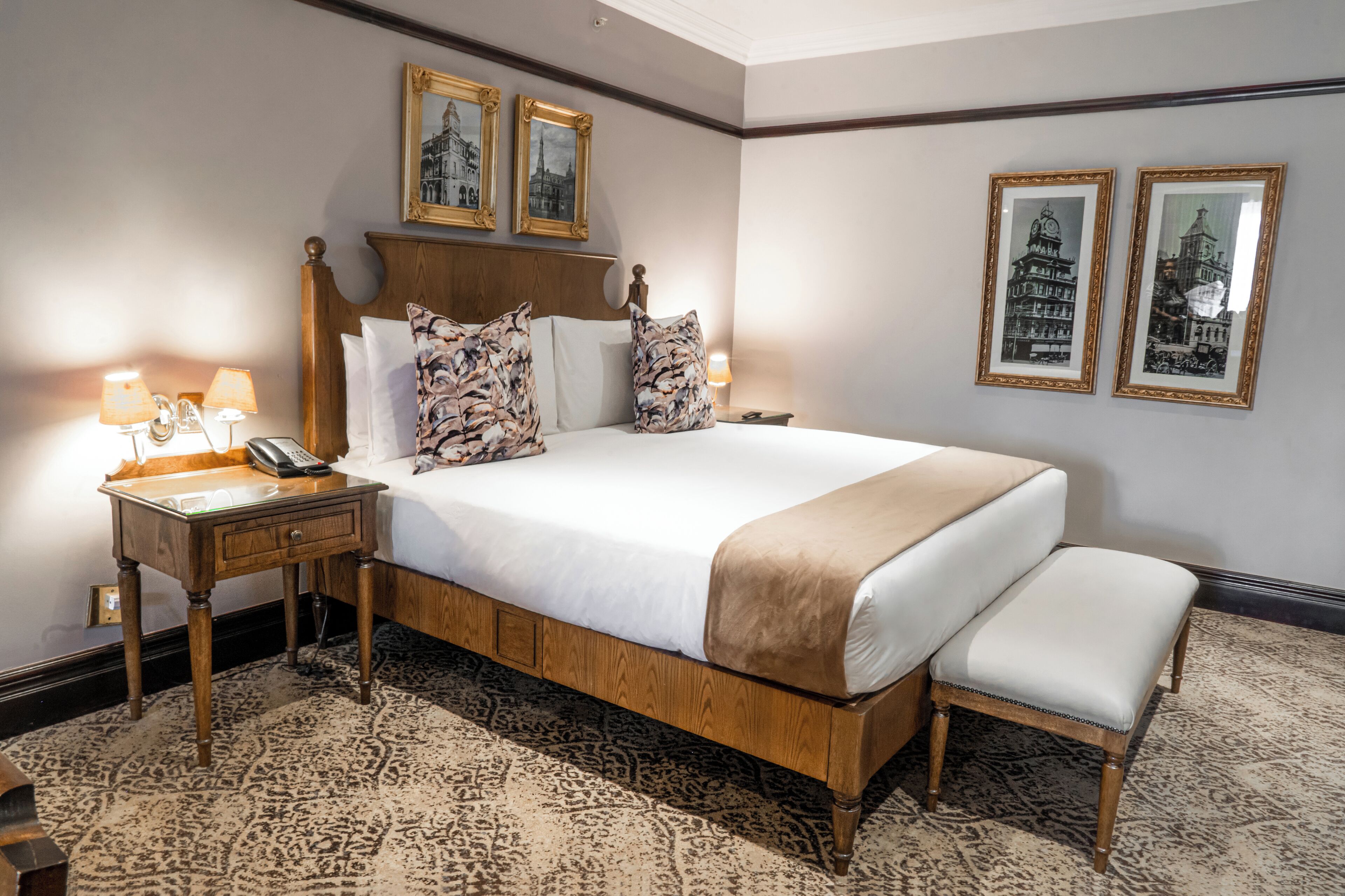 Habitación de hotel con una cama grande, cabecero de madera, dos mesitas de noche con lámparas, teléfono junto a la cama y alfombra estampada.