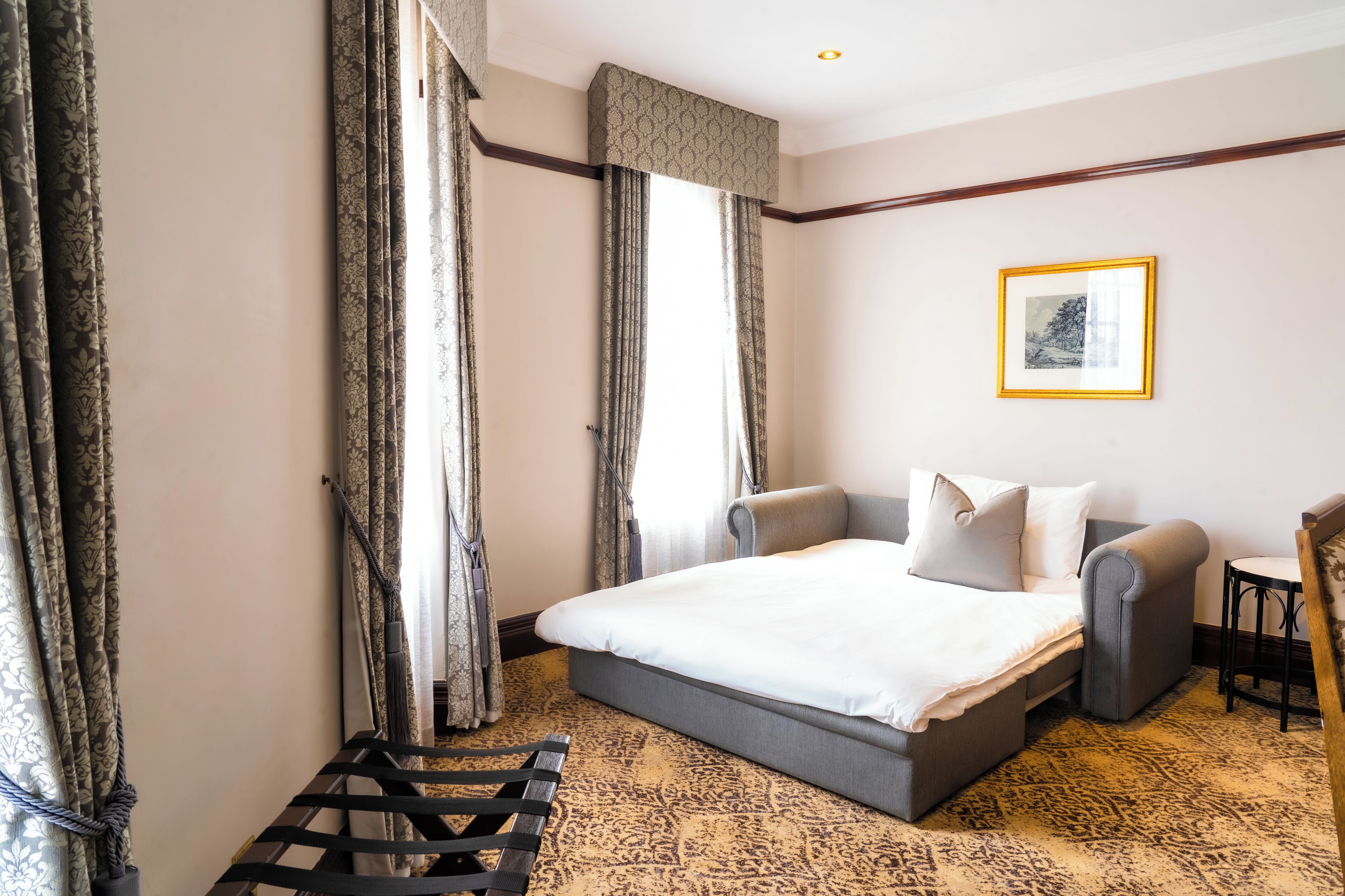 A hotel room with a large bed, a sofa, a small table, and a chair. There are curtains on the windows, and a framed picture on the wall.