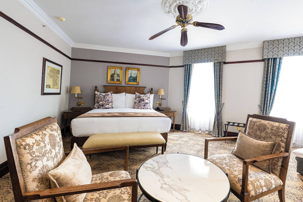 A hotel room with a large bed, two armchairs, a small table, and a ceiling fan.