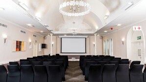 Un salón de conferencias con filas de sillas negras, una pantalla grande, una araña de cristal y un proyector.