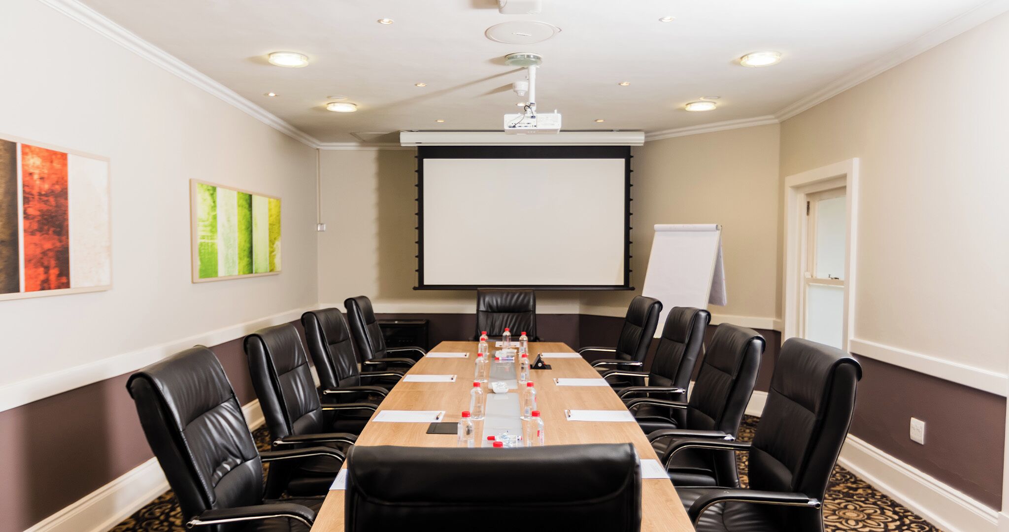 Un salón de conferencias con una larga mesa, sillas de cuero negro, una pantalla de proyector y una pizarra.