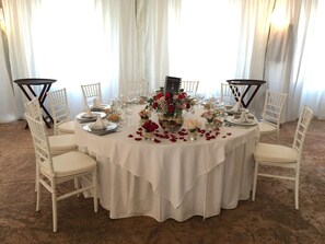 Una mesa redonda dispuesta para un evento formal, con sillas blancas, un mantel blanco y centros de mesa florales.