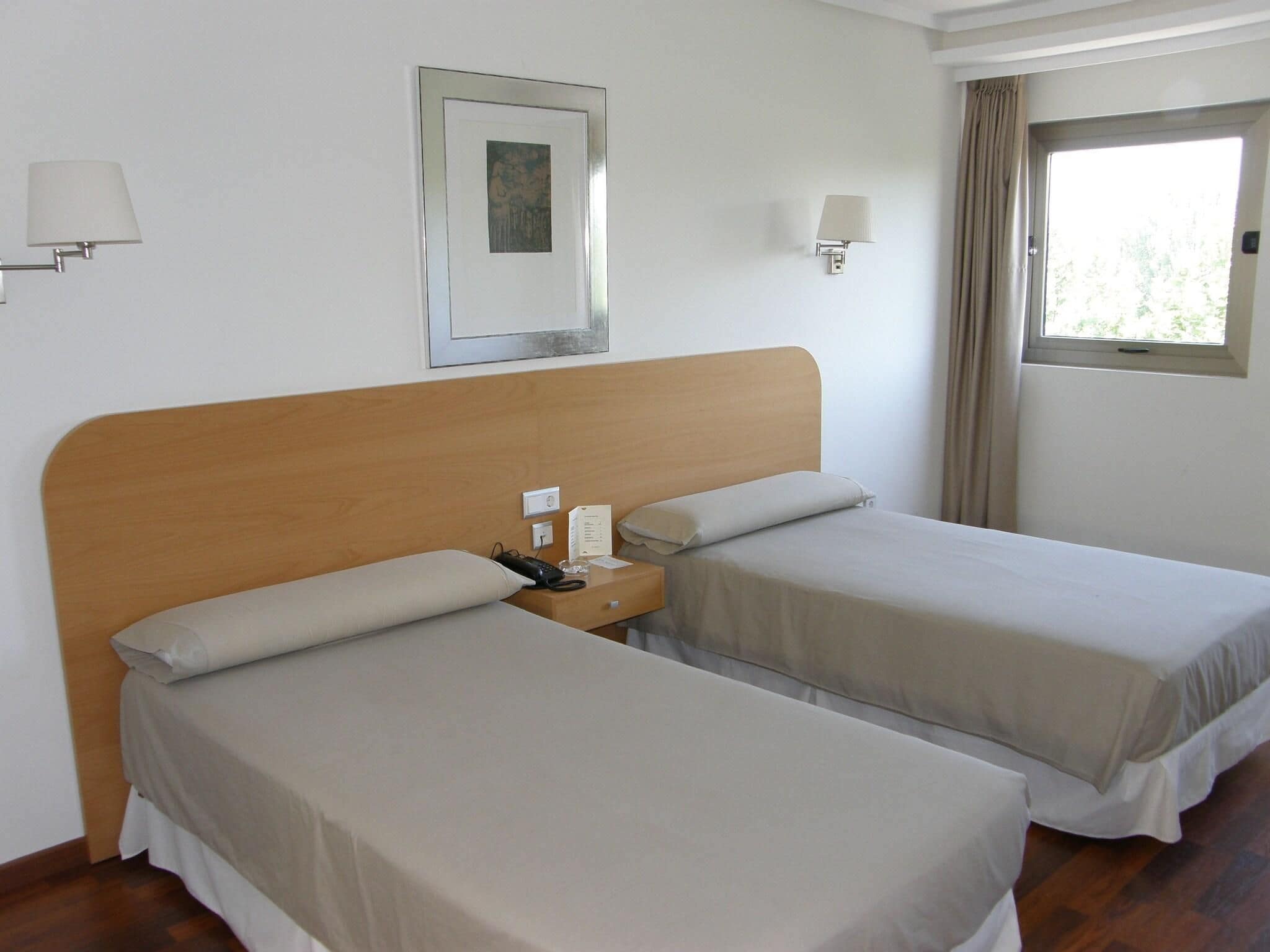 Habitación de hotel con dos camas individuales, cabecero de madera, lámparas de noche, un cuadro enmarcado en la pared y una ventana con cortinas.