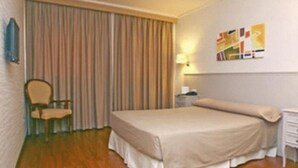 A hotel room with a bed, a chair, a nightstand, a lamp, and a curtain.