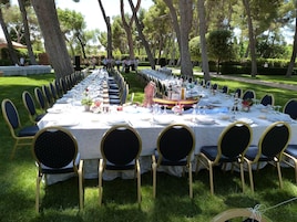 Una larga mesa de comedor al aire libre con sillas, un centro de mesa decorativo con un barco y un césped bien cuidado.