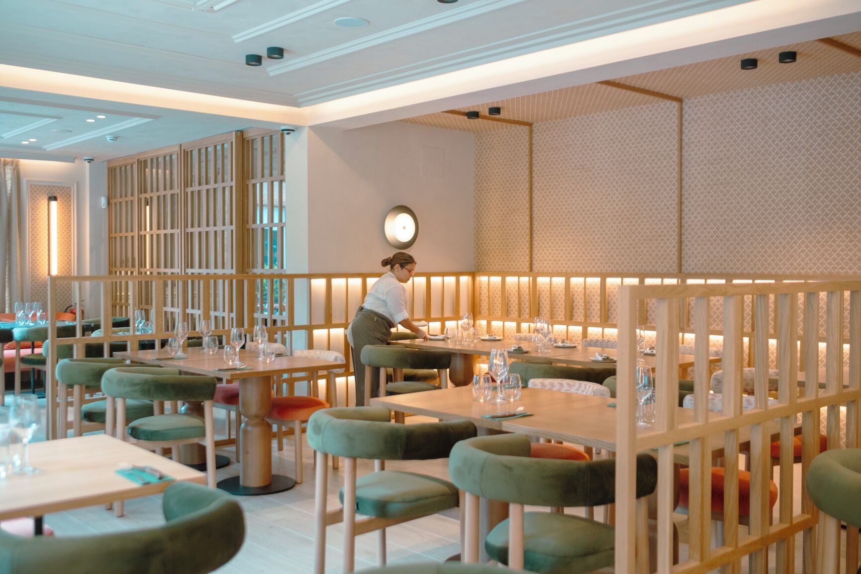 A restaurant with wooden partitions, green upholstered chairs, and a waiter attending to a table.